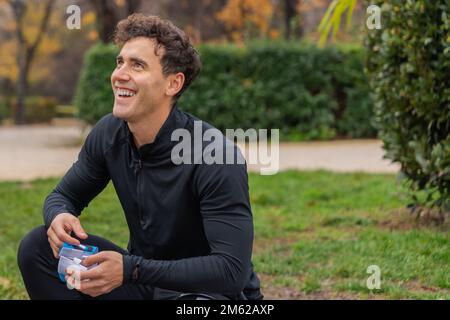 Positive male in casual clothes smiling and looking away while sitting on grass in green park Stock Photo