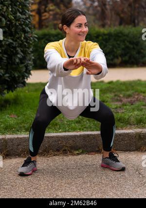 Full body of positive young female runner in colorful sweatshirt doing squats in park while looking away Stock Photo