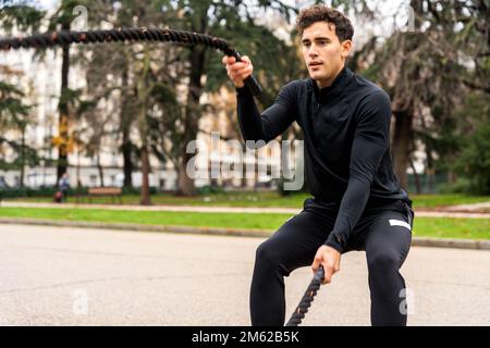Determined young male athlete with short curly hair in black activewear doing exercise with ropes in park with green trees Stock Photo