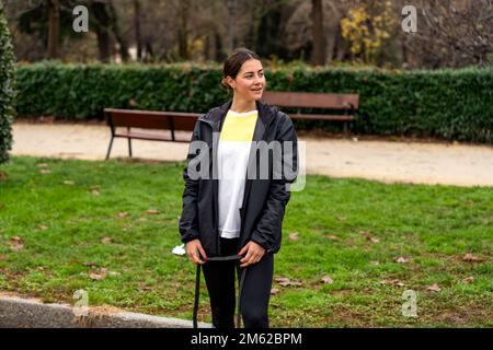 Positive young female athlete in black windbreaker standing with skipping rope in park near benches and looking away Stock Photo