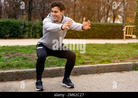 Full body of positive young male runner in sportswear doing squats on asphalt road in park while looking away Stock Photo