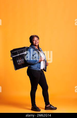 Deliverywoman standing in studio over yellow background carrying takeout thermal backpack ready to deliver meal order to customer. Food transportation service and takeaway concept Stock Photo