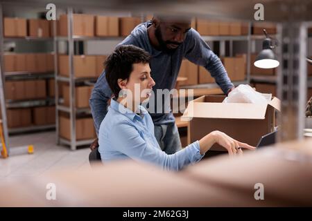 Storehouse employees discussing shipping logistics while looking at cargo stock on laptop computer, preparing packages for delivery. Workers putting order in cardboard box in warehouse Stock Photo