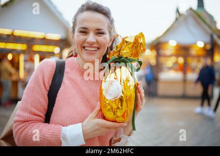 Easter fun. modern middle aged woman in pink blouse with golden easter egg at the fair in the city. Stock Photo