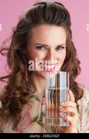 Portrait of smiling stylish 40 years old woman in floral dress with glass of water against pink background. Stock Photo