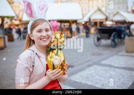 Easter fun. Portrait of happy modern child in pink jacket with golden easter egg at the fair in the city. Stock Photo