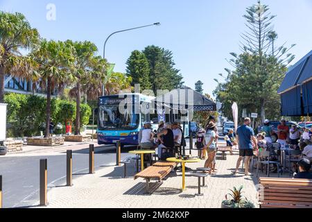 Sydney bus on one way road through Avalon Beach village centre, summers day 2023, past people at a cafe coffee shop,Sydney,Australia Stock Photo