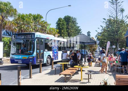 Sydney bus on one way road through Avalon Beach village centre, summers day 2023, past people at a cafe coffee shop,Sydney,Australia Stock Photo