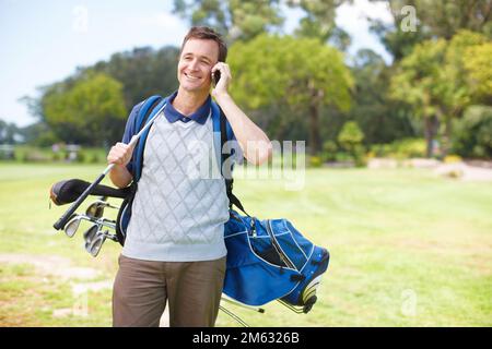 Feeling like joining me for a round. Smiling golfer using his mobile on the course while carrying his clubs. Stock Photo