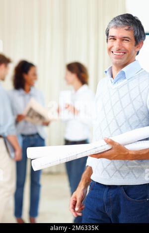 Male architect holding blueprint. Mature male architect holding blueprints with colleagues discussing in background. Stock Photo