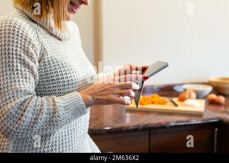 Crop smiling adult lady in cozy sweater tapping screen of tablet while standing against cabinets in kitchen at home Stock Photo