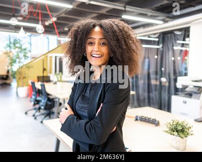 Positive African American woman with Afro hairstyle in formal outfit smiling and looking at camera while standing with arms crossed in office Stock Photo