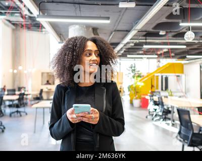 Smiling African American woman in formal wear standing in contemporary workspace and browsing mobile phone looking away Stock Photo
