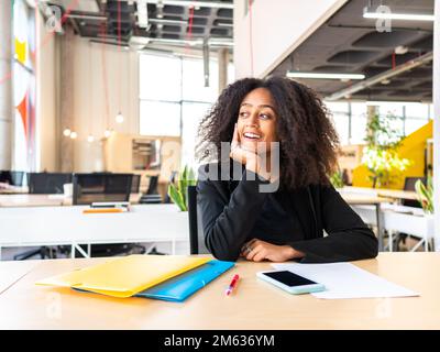 Positive African American businesswoman in formal wear sitting at table leaning on hand during workday looking away dreamily Stock Photo