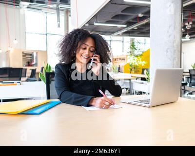 Positive young African American female entrepreneur in formal outfit sitting at table with laptop having phone conversation while writing down informa Stock Photo