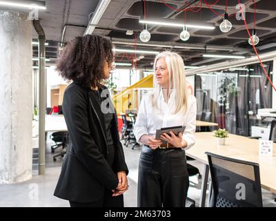 Senior executive in formal clothes standing in modern office with tablet while talking to black female employee during workday Stock Photo