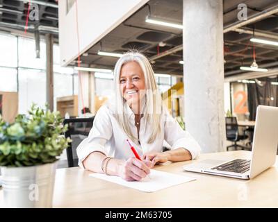 Positive aged female manager in formal outfit sitting at table writing notes on paper while working on netbook in contemporary workspace looking at ca Stock Photo
