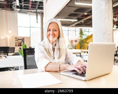 Positive senior female manager in formal clothes sitting at table typing information on netbook during work in modern workspace Stock Photo