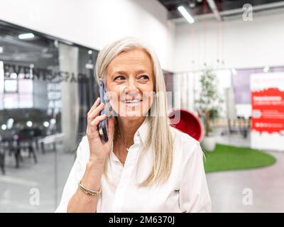 Positive elderly female boss in white shirt speaking on cellphone having business call during work in modern company Stock Photo