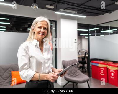 Positive elderly female manager in classy wear smiling and looking at camera while browsing tablet during work in modern office Stock Photo