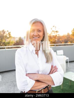 Positive elderly female manager in classy clothes smiling and looking at camera while standing in backlit with crossed arms Stock Photo