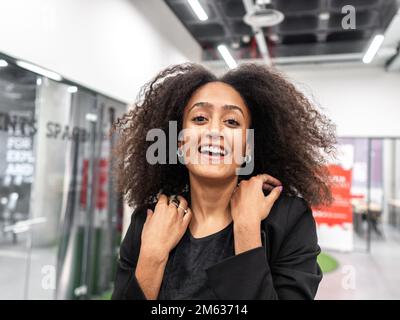 Positive African American young woman with dark Afro hair in classy outfit smiling and looking at camera while standing in corridor of contemporary co Stock Photo