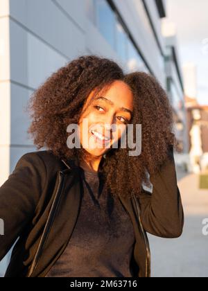 Positive young African American woman with Afro hairstyle standing against sunlight on street near office building looking away Stock Photo