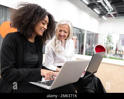Positive young African American female with Afro hairstyle sitting on bench in office typing on netbook while aged executive using computer Stock Photo