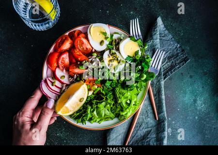 From above cropped unrecognizable hand of woman holding bowl with healthy raw vegetable salad with seeds and boiled eggs on concrete table Stock Photo