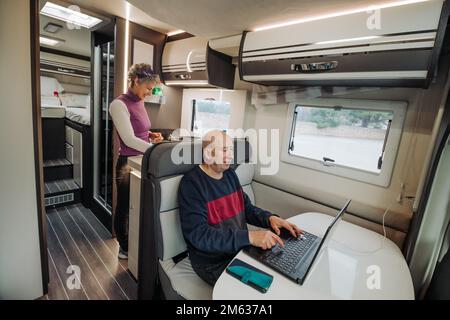 High angle of elderly man in casual clothes sitting at table typing information on netbook while mature woman preparing dinner during travelling in ca Stock Photo