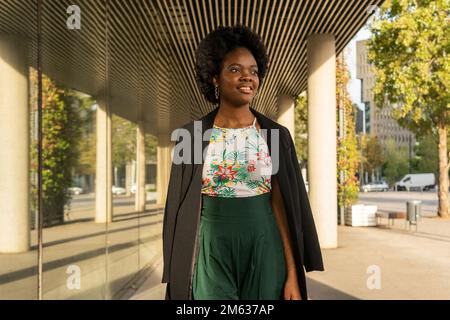 Positive young African American female in black jacket standing near building and looking away thoughtfully on sunny day Stock Photo
