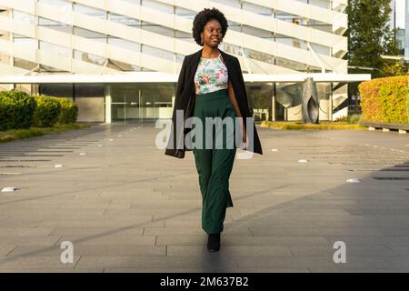 Pensive young African American female in black jacket walking near building and looking away thoughtfully on sunny day Stock Photo