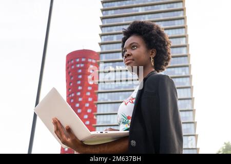 Low angle side view of pensive African American female freelancer with Afro hairstyle in black jacket standing on city street near multistory building Stock Photo