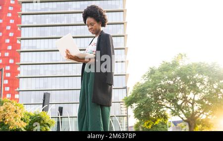 Low angle side view of pensive African American female freelancer with Afro hairstyle in black jacket standing on city street near multistory building Stock Photo