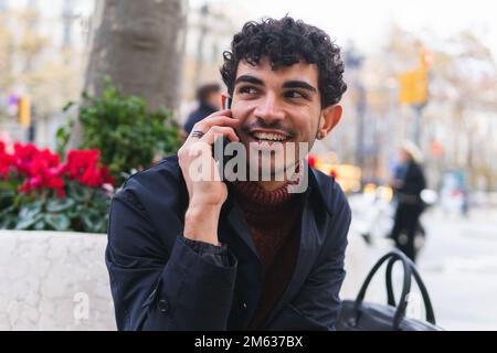 Positive Hispanic man looking away while speaking on mobile phone against blurred background of autumn park Stock Photo