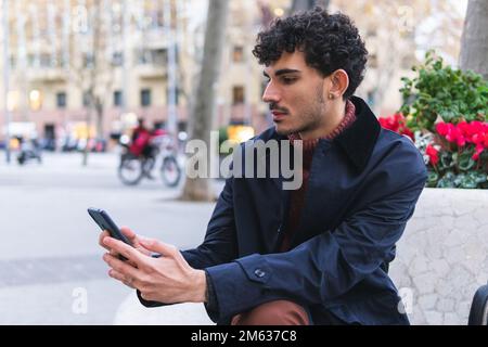 Focused ethnic male in black jacket sitting on bench on city street and browsing smartphone Stock Photo