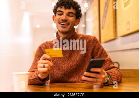 Cheerful man in sweater with credit card sitting at wooden table in cafe having paper cup of hot beverage and using cellphone Stock Photo