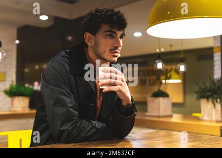 Young male in warm clothes sitting at round table in modern cafeteria while having paper cup of hot beverage Stock Photo
