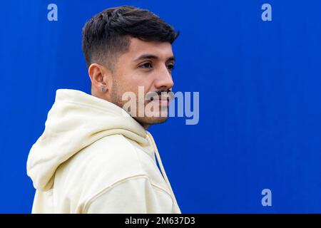 Young Hispanic male in white hoodie and mustache looking away against blue wall on city street Stock Photo