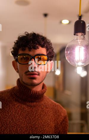 Young male in warm sweater and sunglasses standing near transparent glass light bulb in modern cafe Stock Photo