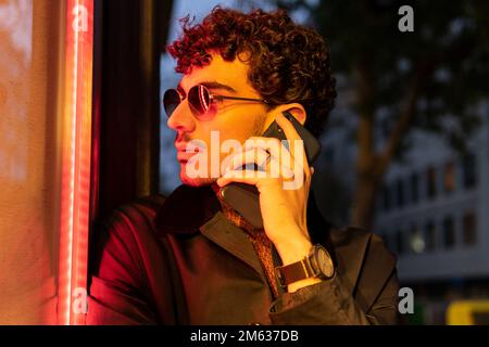 Young male with curly hair in stylish outfit and sunglasses talking phone while standing near window on street in evening Stock Photo