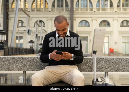Positive African American male in smart casual outfit sitting on bench with suitcase and messaging via mobile phone while waiting for train Stock Photo