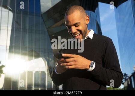 From below of cheerful African American businessman in smart casual outfit chatting on mobile phone while standing against modern business center Stock Photo