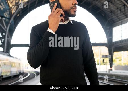 Low angle of cropped young black man in smart casual clothes speaking on cellphone while waiting for train arrival Stock Photo