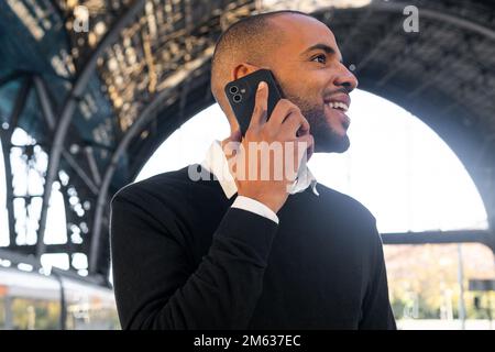Low angle of positive young black man in smart casual clothes speaking on cellphone while waiting for train arrival Stock Photo