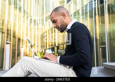 Side view of focused black man in smart casual clothes typing information on netbook while working on business project outdoors Stock Photo