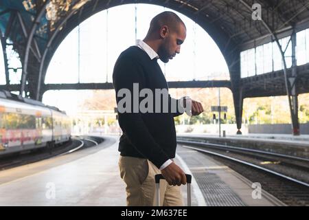 Side view of punctual ethnic man in smart casual clothes with suitcase standing on railway station while waiting for train Stock Photo