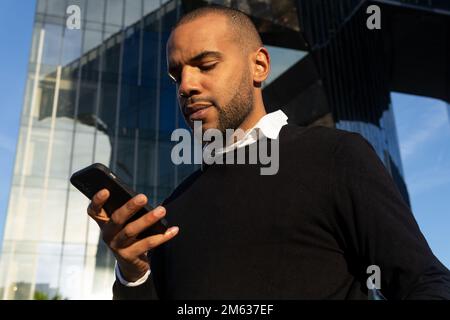 From below of focused African American businessman in smart casual outfit chatting on mobile phone while standing against modern business center Stock Photo