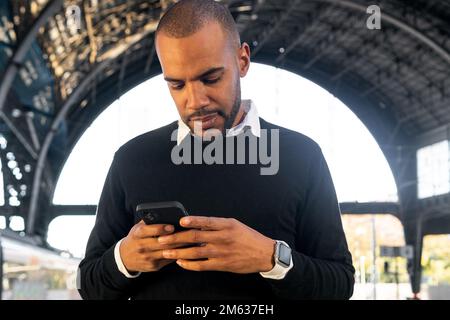 Low angle of positive young black man in smart casual clothes text message on cellphone while waiting for train arrival Stock Photo