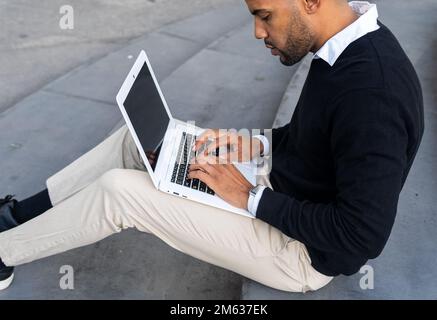 From below focused black man in smart casual clothes typing information on netbook while working on business project outdoors Stock Photo